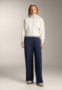 Fată tânără cu păr blond ondulat, îmbrăcată într-un pulover alb cu fermoar și guler, pantaloni largi bleumarin și pantofi bej, pe un fond simplu.