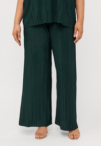 Un mannequin portant un haut ample plissé de couleur vert foncé et un pantalon large assorti, avec des sandales à talons dorés, se tient devant un fond uni.