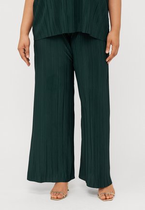 Un mannequin portant un haut ample plissé de couleur vert foncé et un pantalon large assorti, avec des sandales à talons dorés, se tient devant un fond uni.