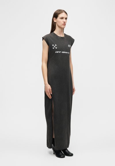 Γυναίκα που στέκεται, φορώντας μακριά μαν sleeveless ρόμπα σε σκούρο γκρι με λογότυπα και πλαϊνή σχισμή, συνδυασμένη με μαύρα μπότες αστραγάλου με τακούνι.