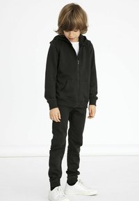 Felpa nera con zip e tasca frontale, abbinata a pantaloni slim-fit coordinati. Sneakers bianche completano l'outfit. Tessuto morbido, design casual.