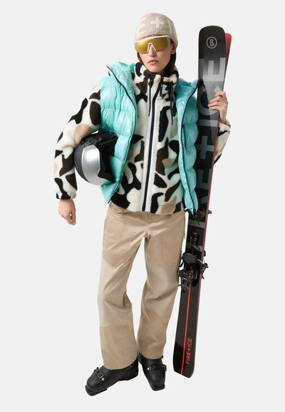 Mintgrüner Puffer-Weste, gemusterte Fleecejacke, beige Schneehose, schwarze Skischuhe, silberner Helm, goldene Sonnenbrille und schwarze Skier mit roten Akzenten.