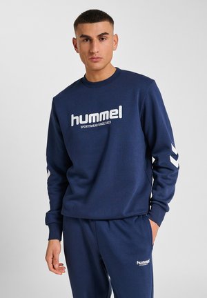 Man in een marineblauwe hummel sweatshirt en bijpassende sweatpants, staand met één hand in de zak tegen een effen grijze achtergrond.