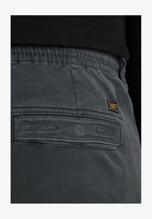 Pantaloni grigi scuri con cintura elastica, caratterizzati da una tasca posteriore con etichetta in tessuto e cuciture a contrasto. Texture complessiva liscia.