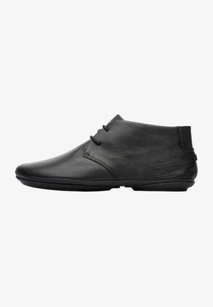 Chaussure noire en cuir à cheville avec une texture lisse, un bout rond, un design minimaliste et un panneau latéral élastique. Les lacets sont fixés à l'avant.