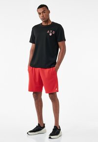 Czarna koszulka sportowa z krótkim rękawem i czerwonymi akcentami, w połączeniu z czerwonymi szortami. Model nosi czarno-białe buty sportowe. Gładki, lekki materiał.