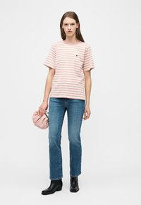 Γυναίκα που φοράει ροζ και λευκή ριγέ μπλούζα, μπλε τζιν, μαύρες μπότες, κρατώντας μια μικρή ροζ τσάντα φάκελο, στέκεται μπροστά σε λευκό φόντο.