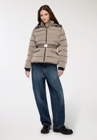 Giubbotto imbottito in taupe chiaro con chiusura a zip, vita elastica e cappuccio. Abbinato a jeans blu a gamba ampia e scarpe nere chunky.