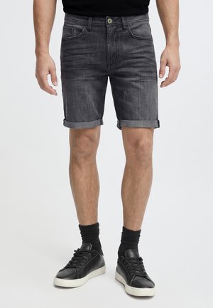 Homme portant un short en denim gris foncé retroussé, des chaussettes noires et des baskets noires, debout devant un fond uni.