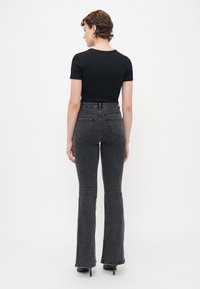 Kvinne med kort brunt hår iført en tettsittende svart kortermet topp, høy midje falmede svarte flagrende jeans og svarte høye sko, stående med ryggen til.