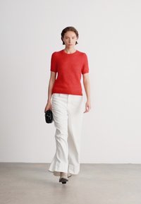 Piros, rövid ujjú kötött pulóver, fehér, bő szabású nadrággal párosítva. Kiegészítve egy kis fekete táskával. Az egyszínű háttér fokozza a figyelmet.