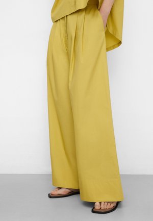 Persoon draagt mosterdgele wijde broekspijpen en een bijpassend bovenstuk met handen in de zakken, gecombineerd met zwarte sandalen met vierkante neus.