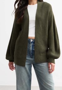 Oliivinvihreä neulottu neuletakki avoimella edustalla, teksturoidulla kankaalla, laskettujen olkapäiden mallilla ja leveillä hihoilla, käytetty valkoisen paidan ja sinisten farkkujen päällä.