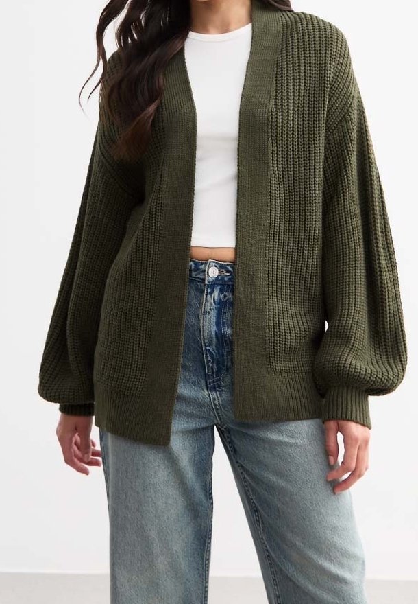 Oliivinvihreä neulottu neuletakki avoimella edustalla, teksturoidulla kankaalla, laskettujen olkapäiden mallilla ja leveillä hihoilla, käytetty valkoisen paidan ja sinisten farkkujen päällä.