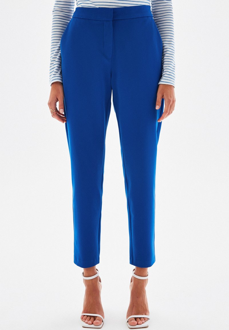 Femme portant un pantalon bleu vif resserré aux chevilles, un haut blanc à rayures longues manches, et des sandales blanches ouvertes, debout sur un fond blanc.