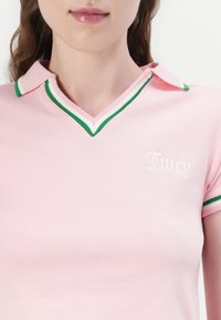 Rožinė polo marškinė su V formos iškirpte, kontrastingais žaliais ir baltais juostelėmis apvadinimu, ir siuvinėtu baltu "Juicy" logotipu krūtinėje.