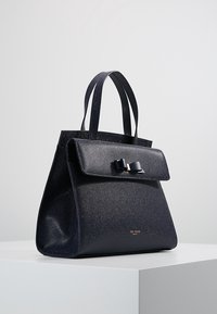 Czarna torebka o teksturowej powierzchni i wyraźnym kształcie, z klapą na froncie i detalem w postaci kokardy, podwójnymi uchwytami oraz subtelnym logo na dole.