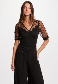 Zwarte blouse met doorzichtige puffmouwen met stippen en ingewikkelde kanten details, gecombineerd met hoge taille zwarte wijde broek.