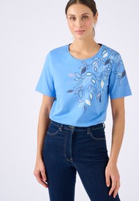 Mujer vistiendo una camisa de manga corta azul claro con un patrón de hojas multicolores en el hombro y unos pantalones vaqueros de tiro alto azul oscuro.