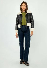 Giacca cropped in pelle nera con foderatura e polsini in sherpa crema, abbinata a un cardigan verde e jeans a vita alta blu scuro. Stivali neri spessi.