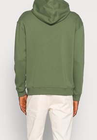 Osoba oblečená v olivově zelené mikině s kapucí a bílých kalhotách, otočená zády, ruce uvolněně podél těla, na jednobarevném pozadí.