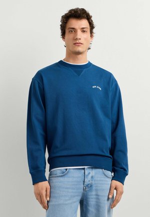 Jonge man met krullend haar draagt een blauwe Pepe Jeans trui en lichtblauwe jeans, staand tegen een effen lichtgrijze achtergrond.