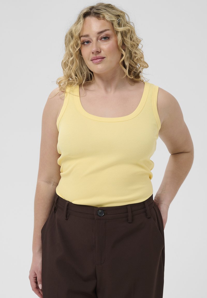 Femme blonde aux cheveux bouclés portant un débardeur jaune et un pantalon marron foncé, debout avec une main dans la poche sur un fond uni.