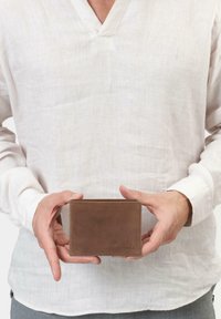 Portefeuille en cuir marron avec une texture lisse, de forme rectangulaire et un subtil détail de couture, tenu à la main contre un fond en tissu clair.