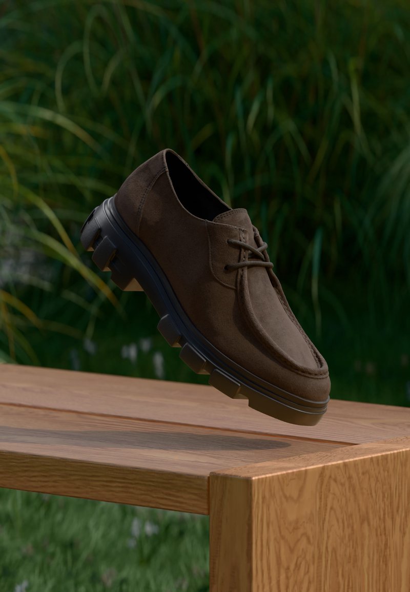 Chaussure en daim marron à lacets avec une semelle noire épaisse suspendue au-dessus d'un banc en bois à l'extérieur avec un fond de feuillage vert.