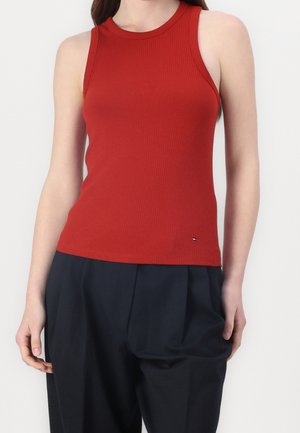 Femme portant un débardeur côtelé rouge sans manches et un pantalon noir ample, debout devant un fond clair uni.