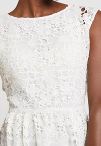 Robe en dentelle blanche avec motifs floraux, col rond et manches courtes. Texture délicate avec des appliqués floraux en superposition.