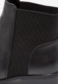 Gros plan sur une botte noire à cheville en cuir lisse avec un panneau latéral élastique côtelé et un talon empilé.