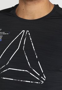 Black athletic T-shirt with a textured finish, featuring a large white graphical logo and small text elements in blue and white.