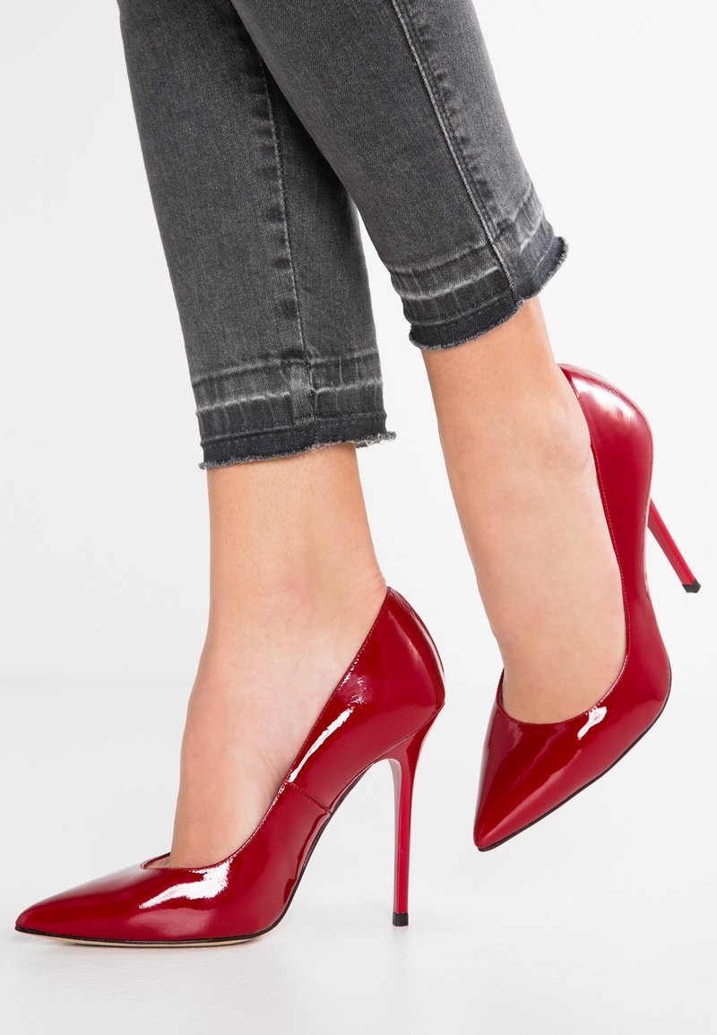 Jambes portant un jean noir coupé associé à des escarpins stilettos à bout pointu rouges brillants sur fond blanc.