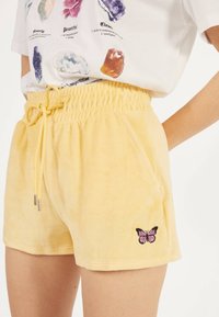 Gula velourshorts med elastisk midja och dekorativ snörning. Har en liten rosa fjärilsbroderi på sidan.