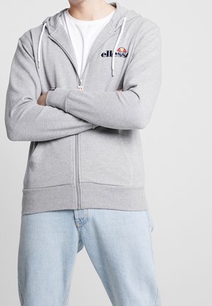 Mann trägt hellgrauen Kapuzenpullover mit Reißverschluss, weißen Kordeln und Logo, weißes T-Shirt und hellblaue Jeans mit verschränkten Armen.