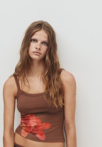 Giovane donna con capelli lunghi e mossi, che indossa una canotta marrone con un disegno di fiori rossi su uno sfondo bianco semplice.