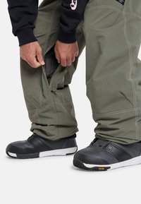 Green snow pants with zipper openings at the ankles. The fabric is durable and smooth, complemented by contrasting white stitching. Black snow boots with Vibram soles.