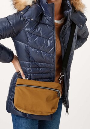 Person trägt eine marineblaue gesteppte Jacke mit Kapuze aus Fell und eine braune Umhängetasche mit vordere Reißverschlusstasche, darunter sind blaue Jeans sichtbar.