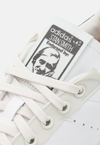 Bílé kožené tenisky s béžovými tkaničkami, s texturovanou jazykovou nálepkou s portrétem a textem "adidas STAN SMITH".