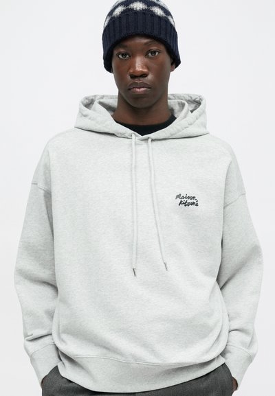 Felpa grigia con cappuccio e un piccolo logo nero sul petto, dotata di cordini e polsini a costine. Indossata con un berretto lavorato a maglia blu navy e bianco.