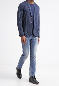 Uomo che cammina, indossa un blazer blu navy, camicia blu scuro, jeans azzurri e scarpe stringate di pelle nere su sfondo bianco.