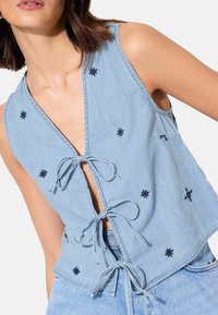 Bezruký světlý denimový top se třemi zavazovacími uzly vpředu a malými tmavými vyšívanými hvězdami, nošený s lehkými džínami ze stejného materiálu.