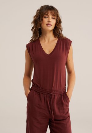 Ženska z rjavimi kodrastimi lasmi, ki nosi brezrokavnik bordo barve z V-izrezom in ujemajoče se hlače z vrvico, stoji z rokami v žepih.