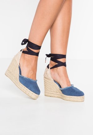 Ben iført blå espadrille-kilehøjhælede sko med flettede rebssåler og sorte bånd omkring anklerne mod en hvid baggrund.
