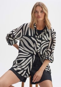 Camicia bianca e nera con righe astratte, tessuto leggero, maniche lunghe, chiusura frontale con bottoni e vestibilità rilassata, abbinata a pantaloni corti scuri.