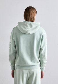 Haut en menthe vert fabriqué en tissu doux, avec une grande capuche, des poignets côtelés et un design imprimé sur la manche gauche.