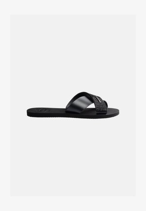 Svart flat hel sandal med brede teksturerte og glatte kryssende remmer på en teksturert såle, vist i sideprofil på hvit bakgrunn.