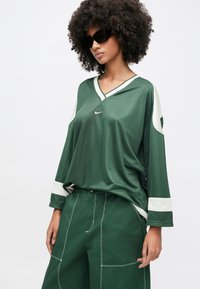 Žena kučeravých vlasov, ktorá má na sebe veľké čierne slnečné okuliare, voľné tmavozelené a biele tričko s dlhým rukávom značky Nike a široké tmavozelené nohavice.