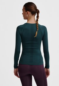 Langemouwen, aansluitend crop top in diep petrol, gemaakt van rekbaar materiaal, voorzien van geribbelde panelen en een naadloos ontwerp, gecombineerd met paarse leggings.
